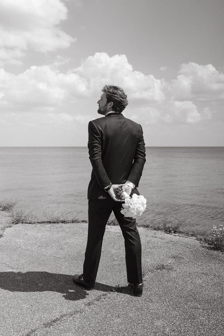 A groom in a black tuxedo standing at the water's edge, looking out over Lake Huron