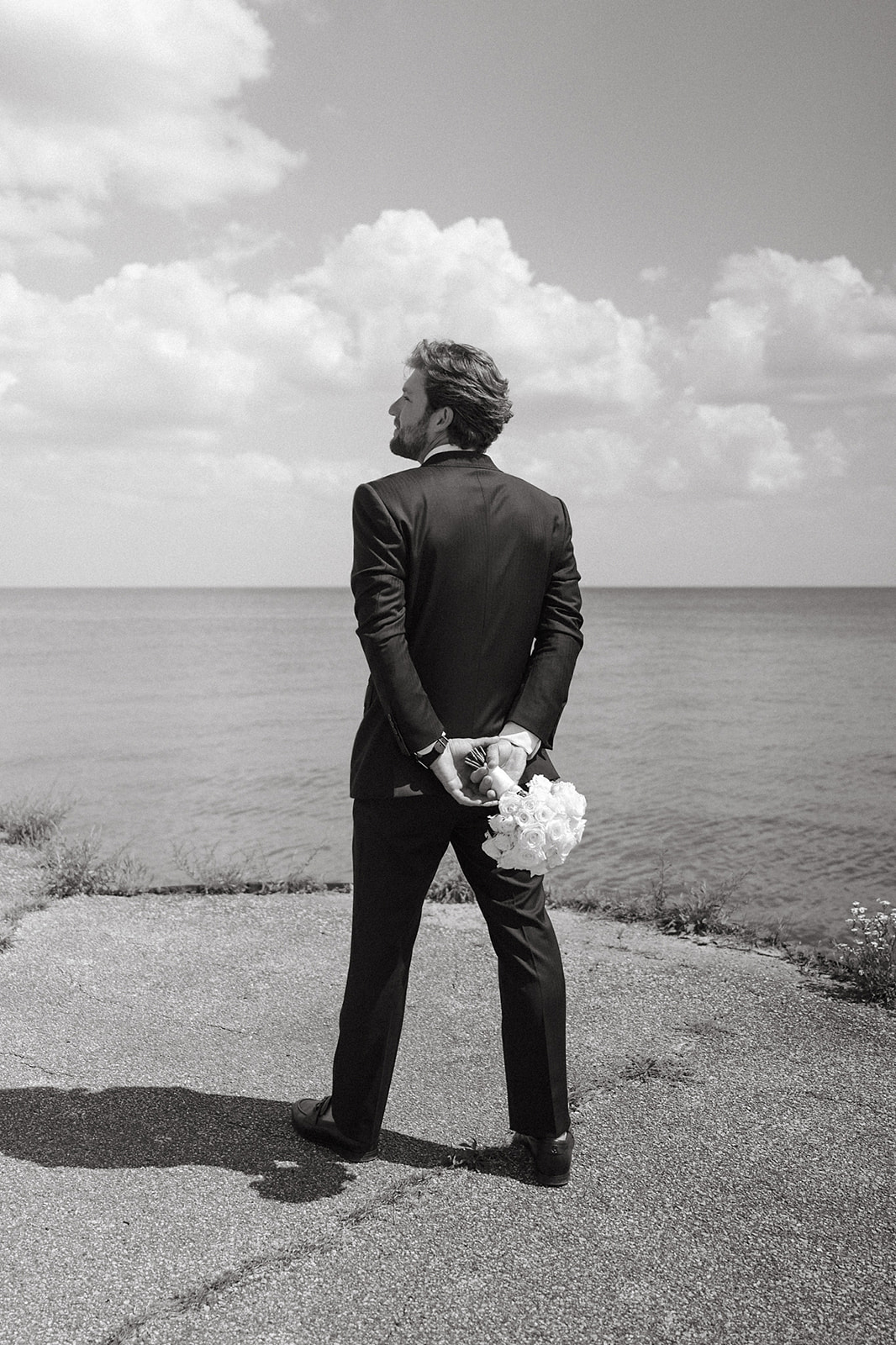 A groom in a black tuxedo standing at the water's edge, looking out over Lake Huron